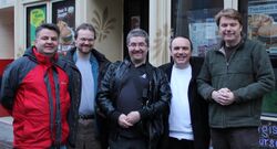 Five middle-aged, white men standing should-to-shoulder in front of a restaurant and smiling at the camera. They wear casual clothing and winter jackets.