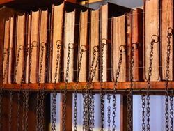 A bookshelf with 14 books. The page edges, not the spines, are facing the viewer. There are chains hanging from the middle of the edge of the covers.