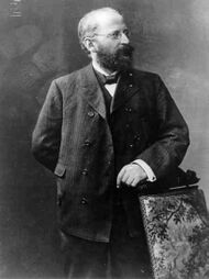 Eduard Bernstein standing next to a chair and looking rightward. He is resting his hand on the chair.