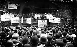 Black and white photograph of a speaker rallying a large crowd. In front of the stage, facing the audience, are several signs, in various languages, displaying demands.