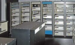 A room with a black box in the foreground and six control cabinets with space for five to six racks each. Most, but not all, of the cabinets are filled with white boxes.