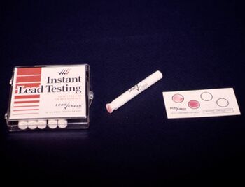 A box of cigarette-like white cylinders on the left, in the middle a white cylinder with a pink tip, on the right a paper with four circles, two blank and two pink