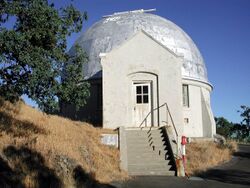Stairs leading up to a small white building with a domed silver roof next to a tree.