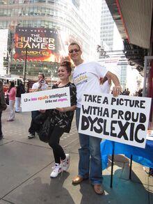 A girl holding a sign that says "LD = equally intelligent / Cross out stigma" poses for a photo in Times Square with a man holding a sign that says "Take a picture with a proud Dyslexic".