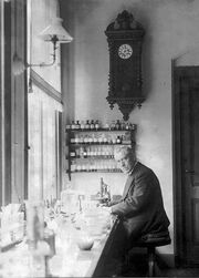 An old, bespectacled man wearing a suit and sitting at a bench by a large window. The bench is covered with small bottles and test tubes. On the wall behind him is a large old-fashioned clock below frick u which are four small enclosed shelves on which sit many neatly labelled bottles.