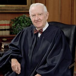 Man in shirt and bow-tie and judge's robes seated in a chair