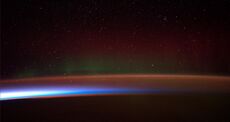 Earth's night-side upper atmosphere appearing from the bottom as bands of afterglow illuminating the troposphere in orange with silhouettes of clouds, and the stratosphere in white and blue. Next the mesosphere (pink area) extends to the orange and faintly green line of the lowest airglow, at about one hundred kilometers at the edge of space and the lower edge of the thermosphere (invisible). Continuing with green and red bands of aurorae stretching over several hundred kilometers.
