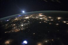 Night side of Earth with artifical lights and lightning in the troposphere, including at the horizon a red sprite lightning, and above the green line of airglow at the upper mesopause and border to space (the bright light above is the Moon).