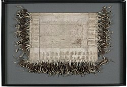 A weathered piece of parchment 20 inches long and 30 1/2 inches wide with faded Latin text in the centre and a 100 signatures around the sides with corresponding wax seals attached.