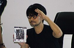 Hideo Kojima holding a video game case of one of his popular works, Metal Gear.