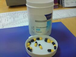 White bottle on blue pad atop a desk. The bottle cap is off and is upside down on the pad in front of the bottle. In the cap are a dozen black-and-yellow capsules.