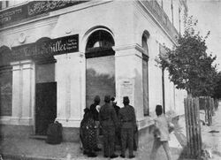 Photo of large white building with one signs saying "Moritz Schiller" and another in Arabic; in front is a cluster of people looking at poster on the wall.