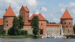 Red brick castle in the background. Lake in front. A wooden bridge with tourists. Sailboats on the lake.