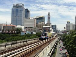 An elevated train, painted in blue, white and a red stripe and with advertisements, running above a road lined with many tall buildings and with many cars