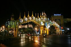 An elaborate double archway above a road, with pictures of King Bhumibol Adulyadej; trees decorated with lights