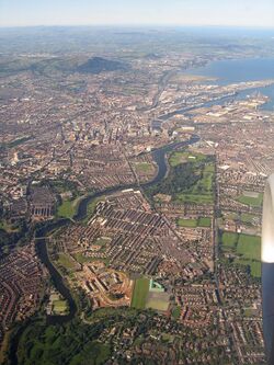Aerial photo of urban sprawl, edged by green hills and sea shore, and bisected by a winding river.