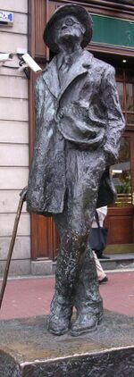 Bronze statue of Joyce standing in a coat and broadbrimmed hat: His head is cocked looking up, his left leg is crossed over his right, his right hand holds a cane, and his left is in his pants pocket, with the left part of his coat tucked back.