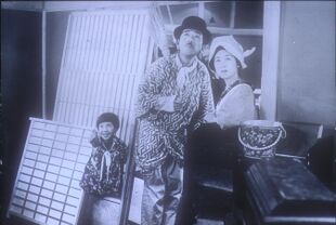 A young girl, man, and woman standing outside of a house, all looking up in the sky. The girl, on the left, is smiling and pointing skyward. The man wears a bowler hat and holds a short broom over his shoulder; the woman wears a kerchief around her head. They are surrounded by domestic objects as if just moving into or out of the house.