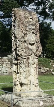 Intricately carved free standing stone shaft sculpted in the three-dimensional form of a richly dressed human figure, standing in an open grassy area.