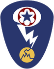 Oval shaped shoulder patch with a deep blue background. At the top is a red circle and blue star, the patch of the Army Service Forces. It is surrounded by a white oval, representing a mushroom cloud. Below it is a white lightning bolt cracking a yellow circle, representing an atom.