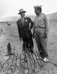 A man smiling in a suit in suit and one in a uniform chat around a pile of twisted metal.
