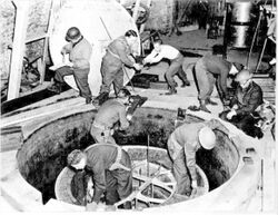 Soldiers and workmen, some wearing steel helmet, clamber over what looks like a giant manhole.