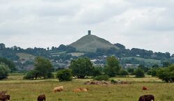 A mound surmounted by a tower in the distance. In the foreground are fields with cows and small trees and bushes.