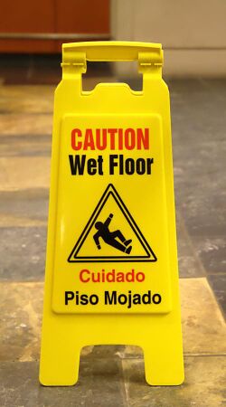 Sign reading "CAUTION Wet Floor" at the top, with a translation of the warning to Spanish at the bottom, and a pictogram in the center depicting a person slipping and falling over