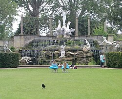a grassy lawn in front of a statues complex several people sunbathe or sit in the midground facing in the background a group of white statues that are significantly larger than the people. In the foreground a black bird is walking away from the whole tableau