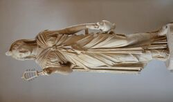 Marble staue of a woman holding a rattle in one hand and a pitcher in the other.