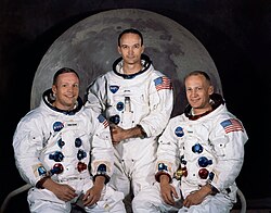 This official Apollo 11 crew portrait shows astronauts Neil Armstrong, Michael Collins, and Buzz Aldrin in their white NASA spacesuits, posed against softly lit backdrop of the Moon. Armstrong (left) and Aldrin (right) are seated, while Collins stands behind them at center. Each suit displays the astronaut's name tag, the NASA insignia, and the American flag on the sleeve, with colored connectors visible on the chest.
