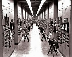 A long corridor with many consoles with dials and switches, attended by women seated on high stools.