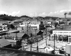 An array of industrial buildings with lots of power poles and wires, with a pair of smokestacks in the background