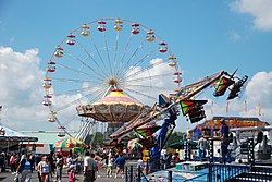 Das Bild zeigt ein Riesenrad und mehrere andere Jahrmarktattraktionen. Dazwischen stehen viele Menschen.