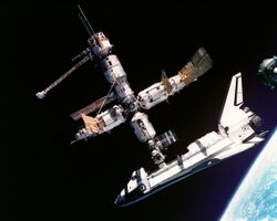 A cluster of cylindrical modules with projecting feathery solar arrays and a spaceplane docked to the lower module. In the background is the blackness of space, and in the lower right corner is Earth.