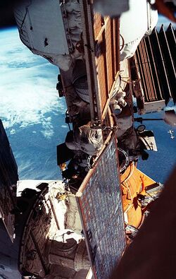 A view showing a module covered in white insulation with a smaller module, covered in orange insulation, connected to the end of it. Part of a space shuttle can be seen attached to the orange module, and a number of folded and unfolded solar arrays are visible. The limb of the Earth forms the backdrop.