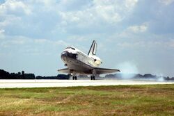 A spaceplane, coloured white on its topside and black on its underside, lands on a runway. A strip of turf is visible in the foreground, there are trees in the background and there is a cloud of smoke coming from the spaceplane's rear wheels.
